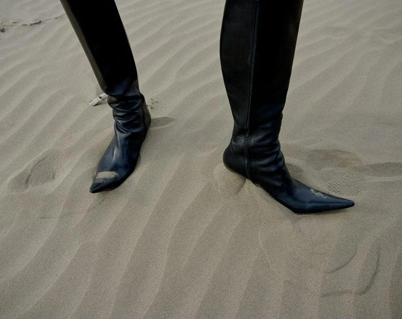 Close-up of a person wearing sleek, black leather knee-high boots standing on a textured sand dune, creating a fashion-forward desert aesthetic.