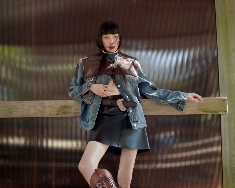 A young Asian woman stands in a fashion pose wearing a denim jacket, leather skirt, brown belt, gold choker, and cowboy boots in a studio.