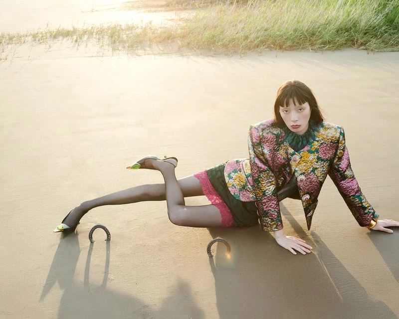 Woman in floral jacket poses on beach wearing heels next to croquet hoops in golden hour light with grassy dunes in the background.