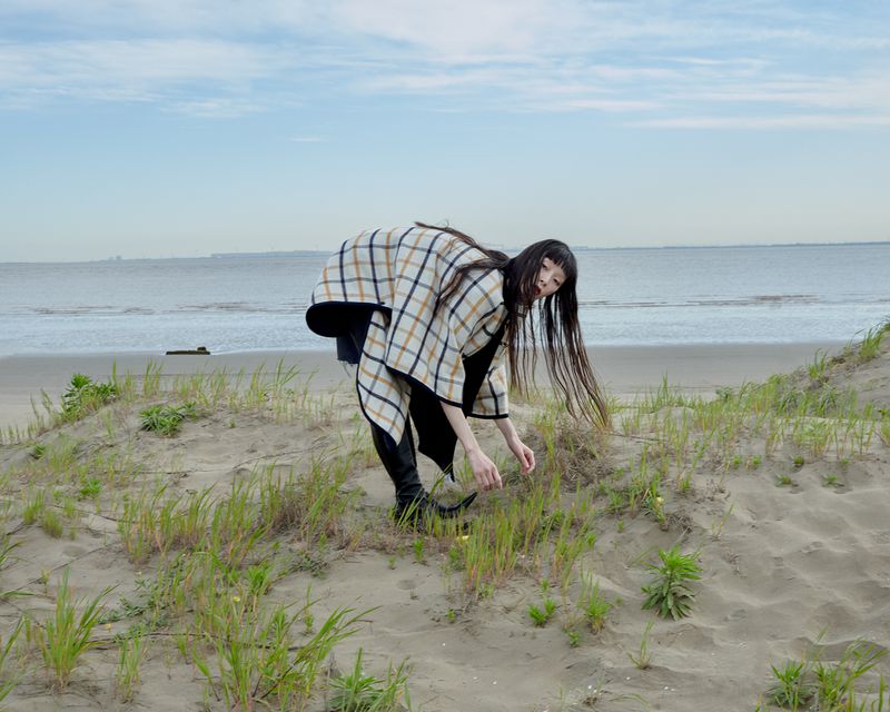 A woman in a patterned coat bends forward to pick something off a sandy dune covered with green grasses near a beach.