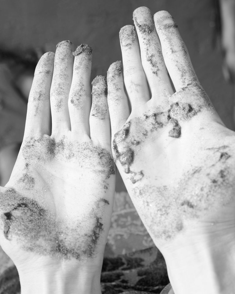 Monochrome image of two open hands with outstretched fingers coated in dark granular material, against a blurry background.
