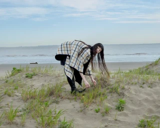 Model in plaid poncho leaning over sand dunes by the ocean for Noblesse Korea