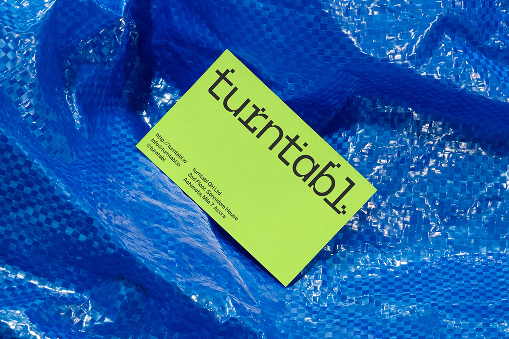 Eye-level shot of a bright green business card with black text, resting on a blue woven plastic background. The card is angled to the left.