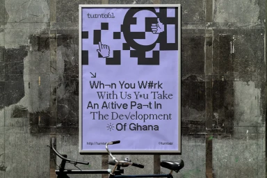 Framed purple poster with pixelated black graphics and text mounted on a weathered concrete wall above bicycles.
