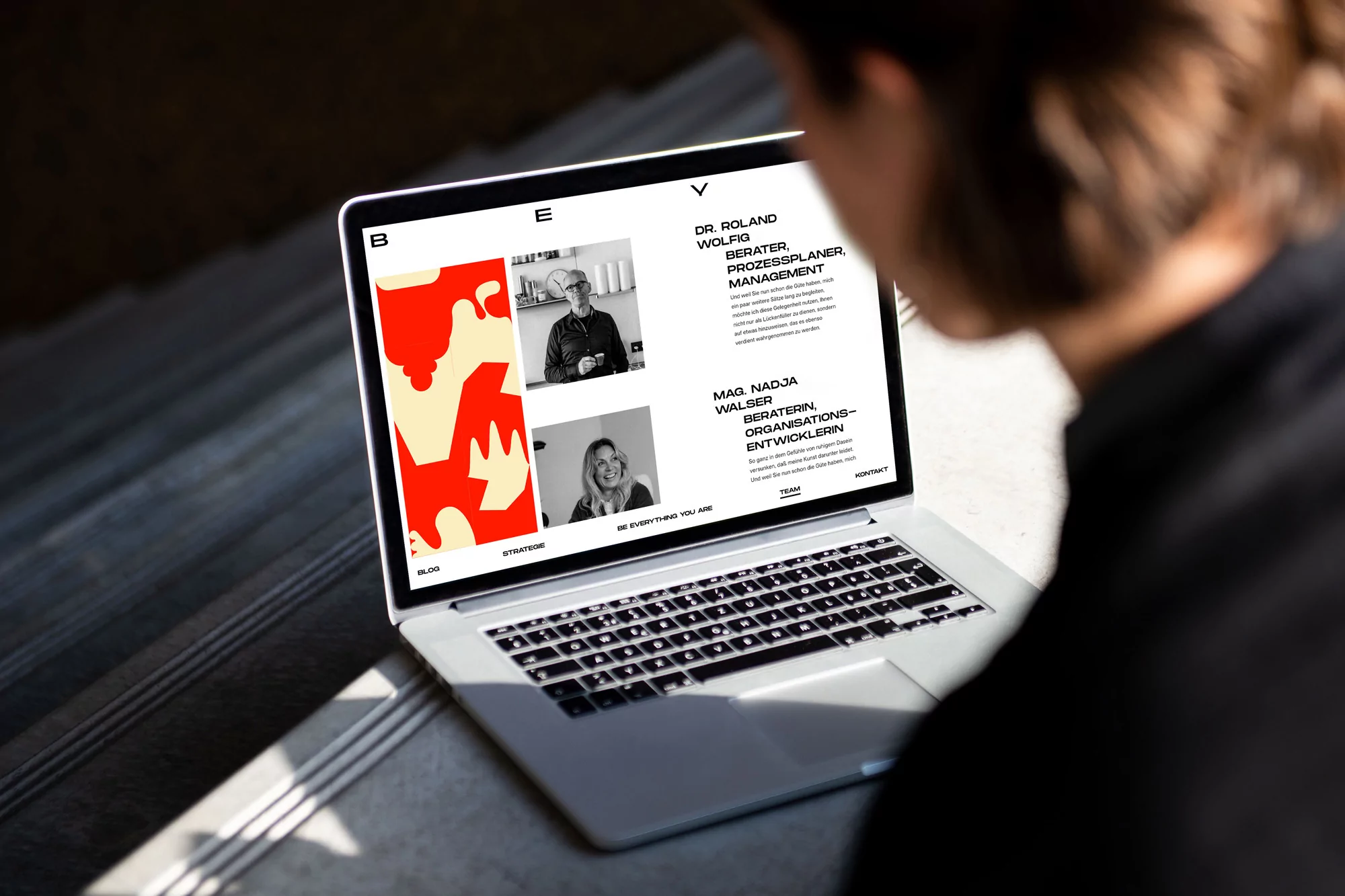 Person views a website on a laptop displaying consultant profiles and abstract design. Sunlight highlights the scene indoors.
