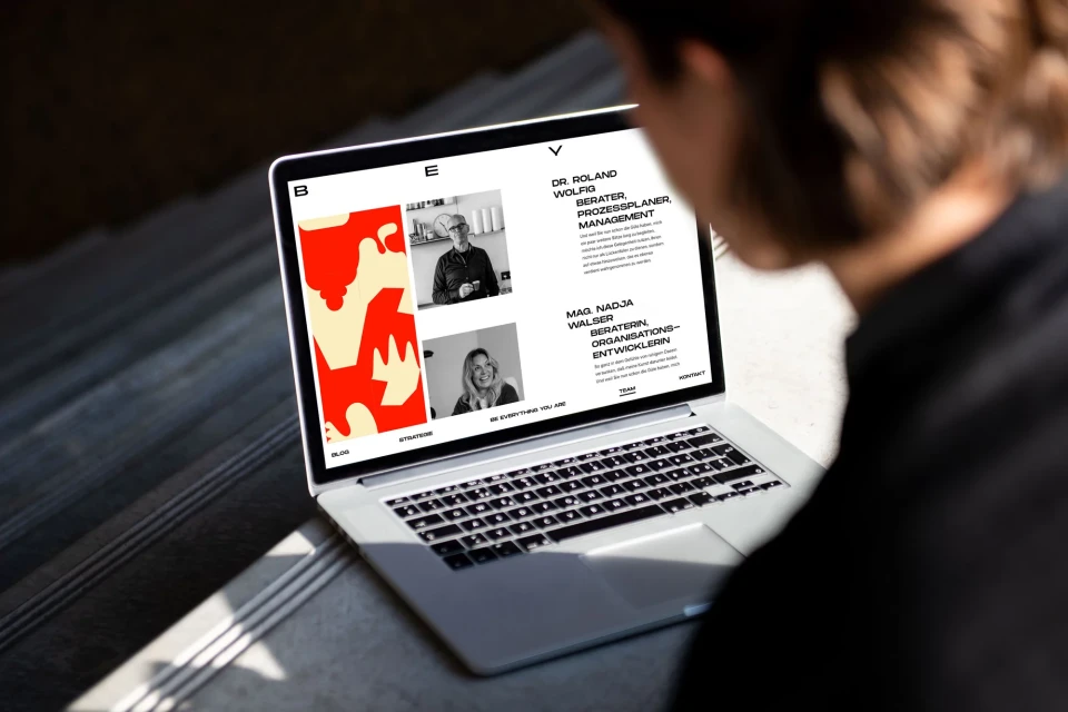 Person views a website on a laptop displaying consultant profiles and abstract design. Sunlight highlights the scene indoors.