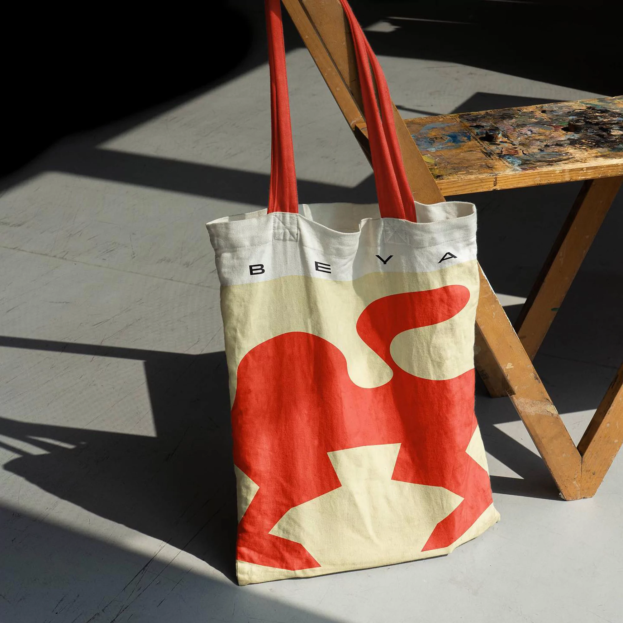 Tote bag with red graphic print and red straps. Shot indoors, light and shadow create strong geometric shapes on the floor.