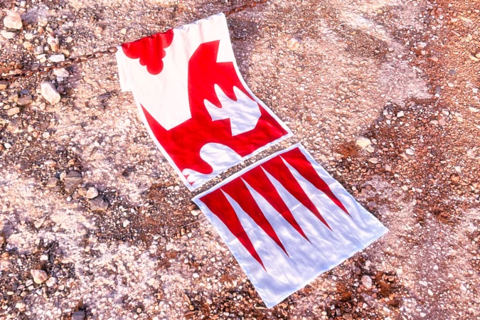 Torn white banner with red graphic of flames and arrowheads lying on a gravel surface in an outdoor setting.