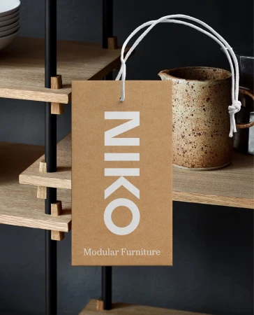 Close-up of a brown rectangular tag reading "Niko Modular Furniture" hanging on black modular shelving, featuring a ceramic vessel.