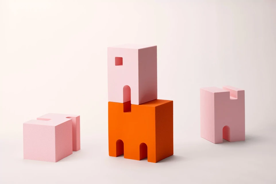 Abstract geometric still life featuring pink and orange building-block shapes against a bright, neutral backdrop in a studio setting.
