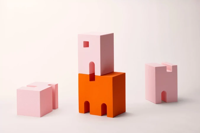 Abstract geometric still life featuring pink and orange building-block shapes against a bright, neutral backdrop in a studio setting.