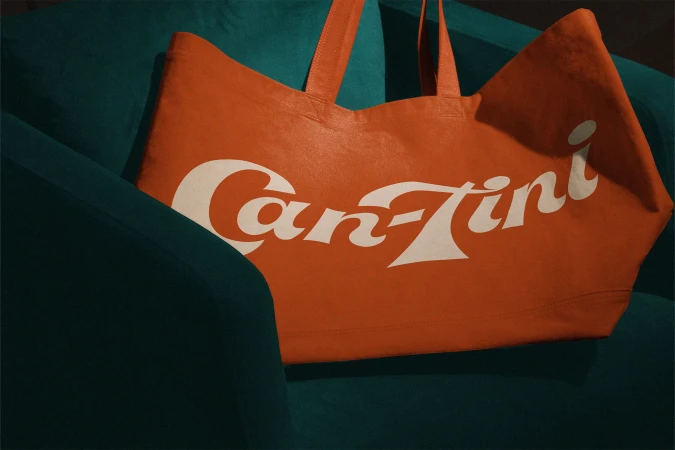 Orange tote bag with the text 'Can-Tini' in white font, placed on a dark green armchair, showcasing a blend of fashion and interior design.