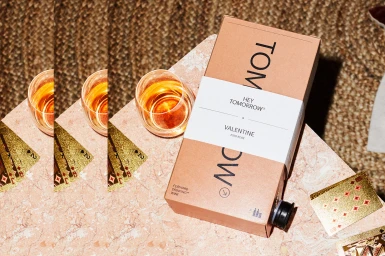 Hey Tomorrow wine box with a glass of amber liquid and gold playing cards on a pink marble tabletop