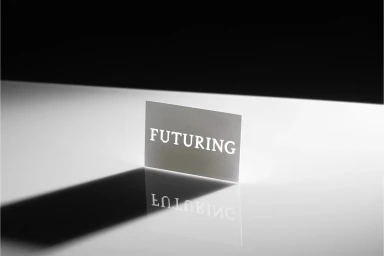 A business card with the text FUTURING standing upright on a reflective white surface with a long shadow.