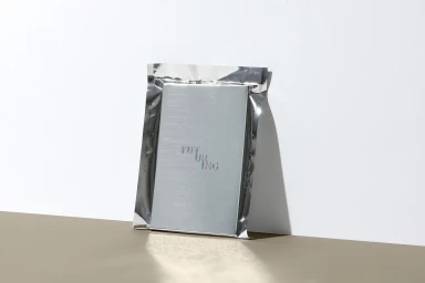 A silver metallic foil packaging pouch resting against a white studio wall with a tan surface below.