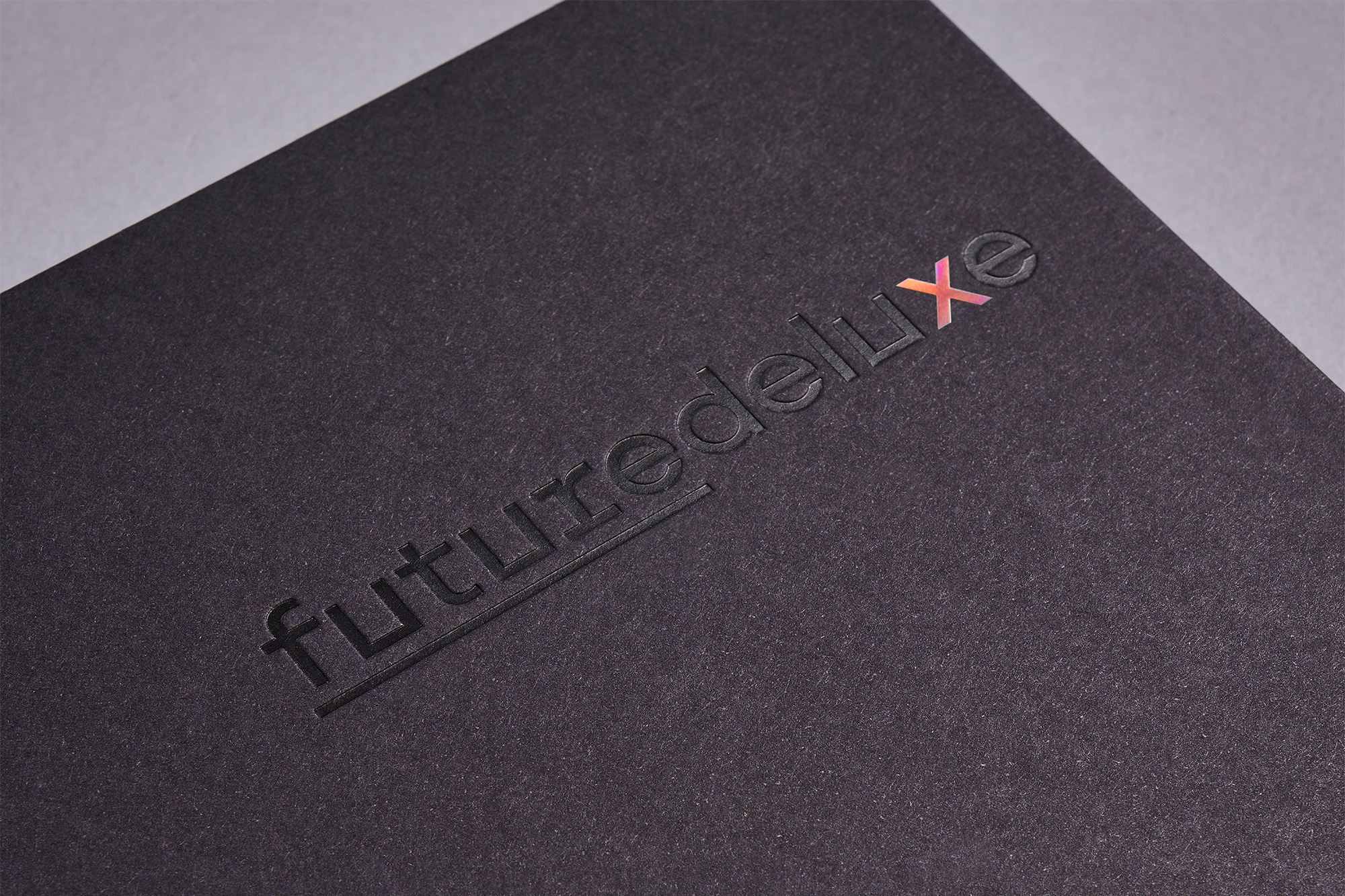 Close-up of a textured black surface with the debossed word "futuredeluxe", partially iridescent. Dark, minimalist design.