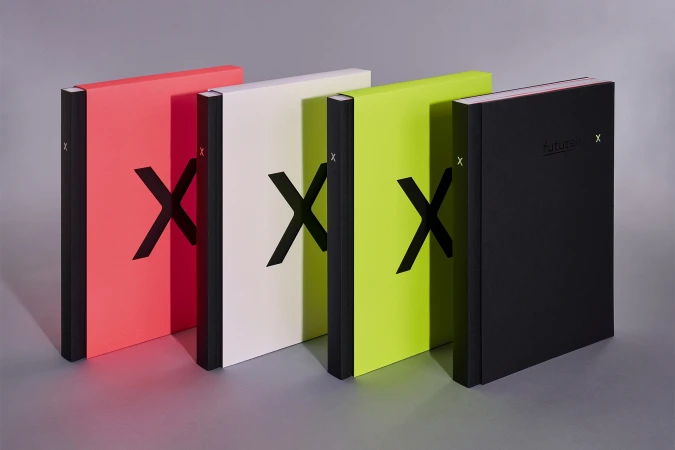 Four colorful books with black 'X' designs stand on a gray surface. The books are coral, off-white, chartreuse, and black.
