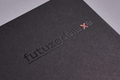 Close-up of debossed Futuredeluxe brand identity on dark grey textured paper packaging set against a pale background