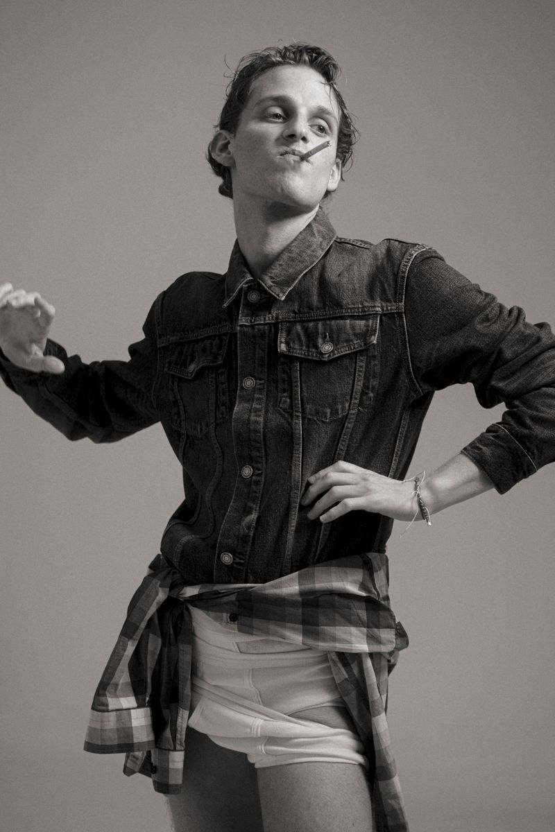 Black and white portrait of a slender adult male with a cigarette in his mouth, wearing a denim jacket and shorts in a photo studio.