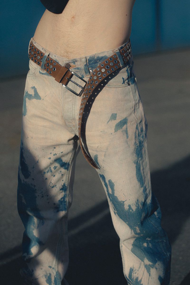 Close-up of a person wearing light-wash jeans with blue dye spots and a studded brown leather belt, showcasing fashion and style.
