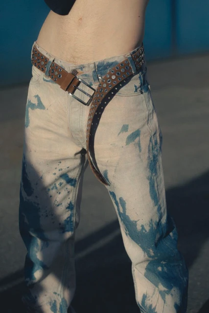 Close-up of a person wearing light-wash jeans with blue dye spots and a studded brown leather belt, showcasing fashion and style.