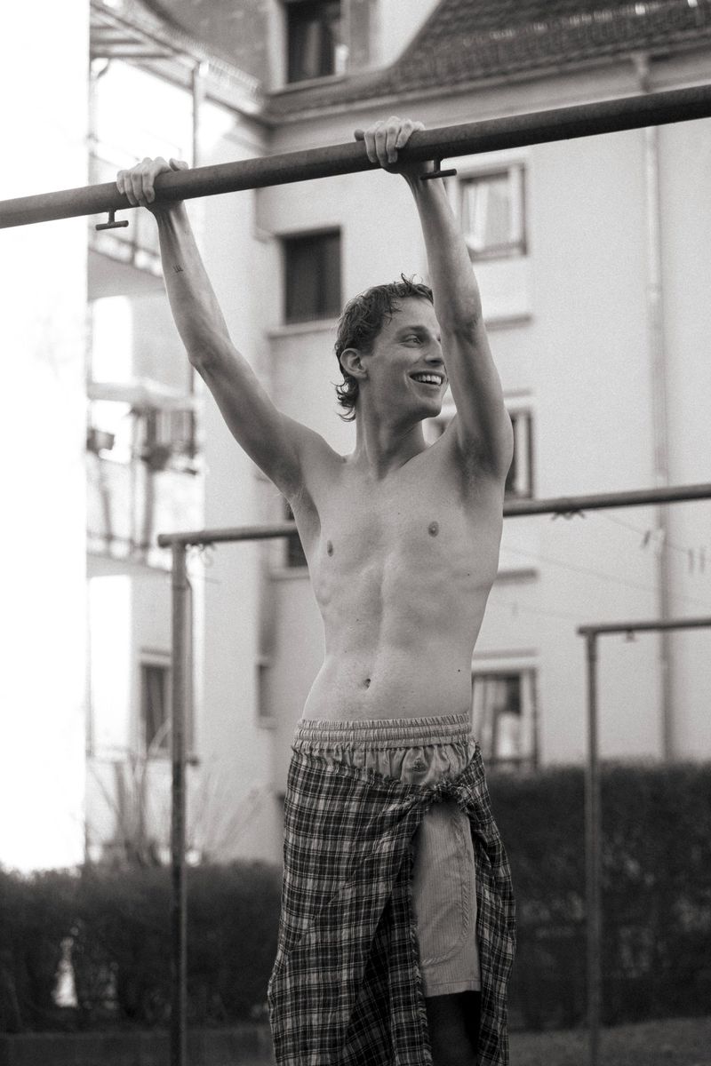 A shirtless adult male is hanging from a metal bar outside in a black and white medium shot with buildings in the background.
