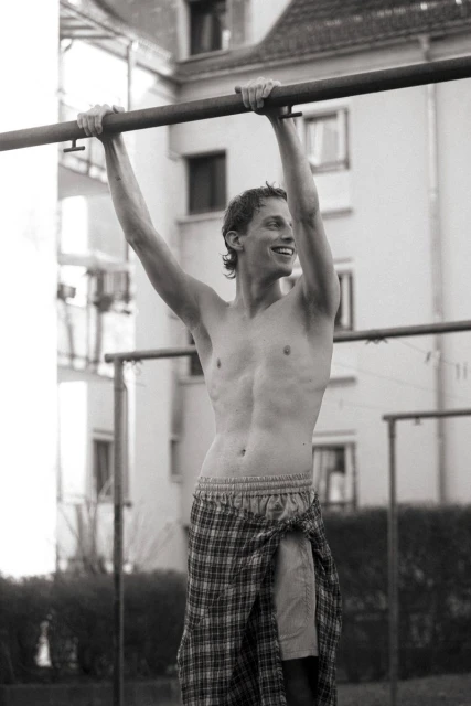 A shirtless adult male is hanging from a metal bar outside in a black and white medium shot with buildings in the background.