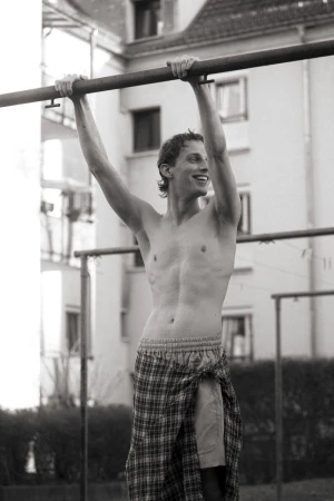 A shirtless adult male is hanging from a metal bar outside in a black and white medium shot with buildings in the background.