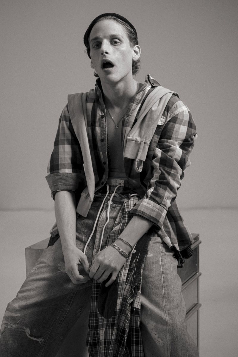 Monochrome shot of a male model sitting on a wooden box, wearing a plaid shirt with a black beanie, looking into the camera.