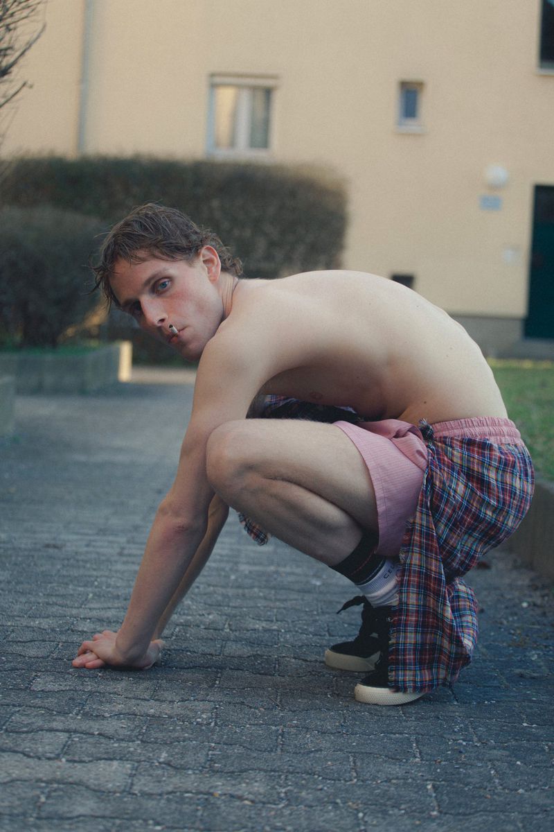 Bare-chested adult male in squatting pose outdoors, wearing casual shorts and sneakers in an urban setting, looking towards the camera.