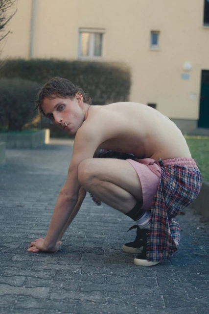 Bare-chested adult male in squatting pose outdoors, wearing casual shorts and sneakers in an urban setting, looking towards the camera.