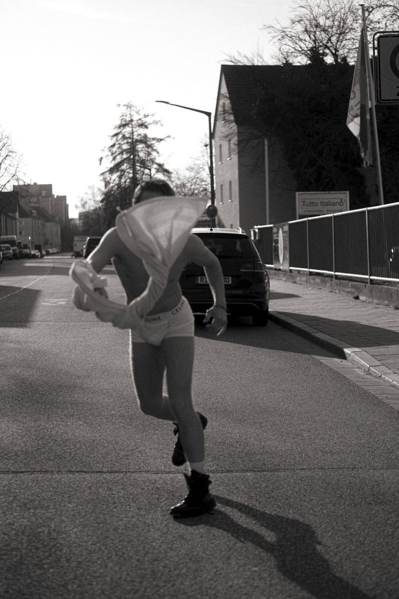 A person wearing underwear and boots runs across a street in a monochrome photo with buildings and cars in the background.
