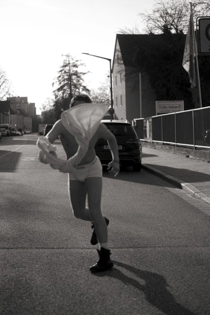 A person wearing underwear and boots runs across a street in a monochrome photo with buildings and cars in the background.
