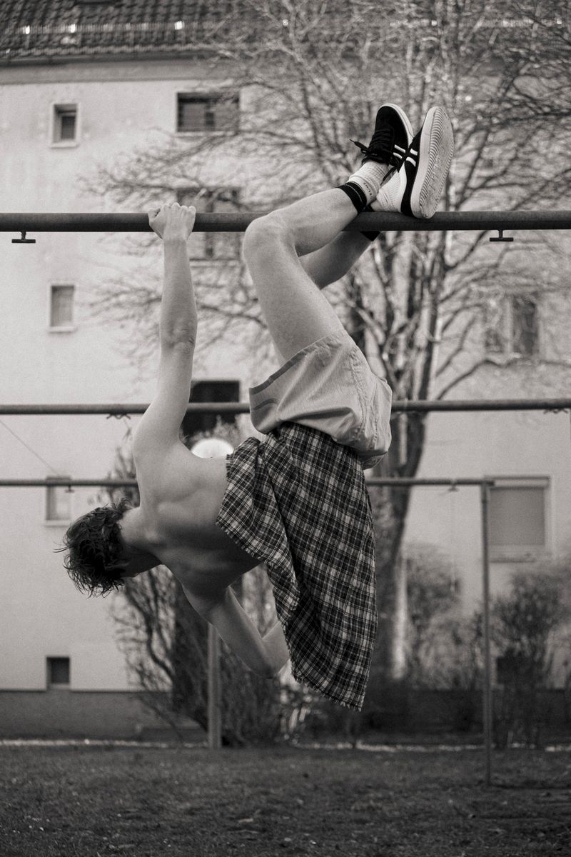 Monochrome image of an adult male hanging upside down from a bar in an outdoor setting with a building and trees in the background.