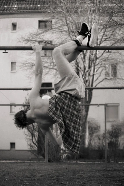 Monochrome image of an adult male hanging upside down from a bar in an outdoor setting with a building and trees in the background.
