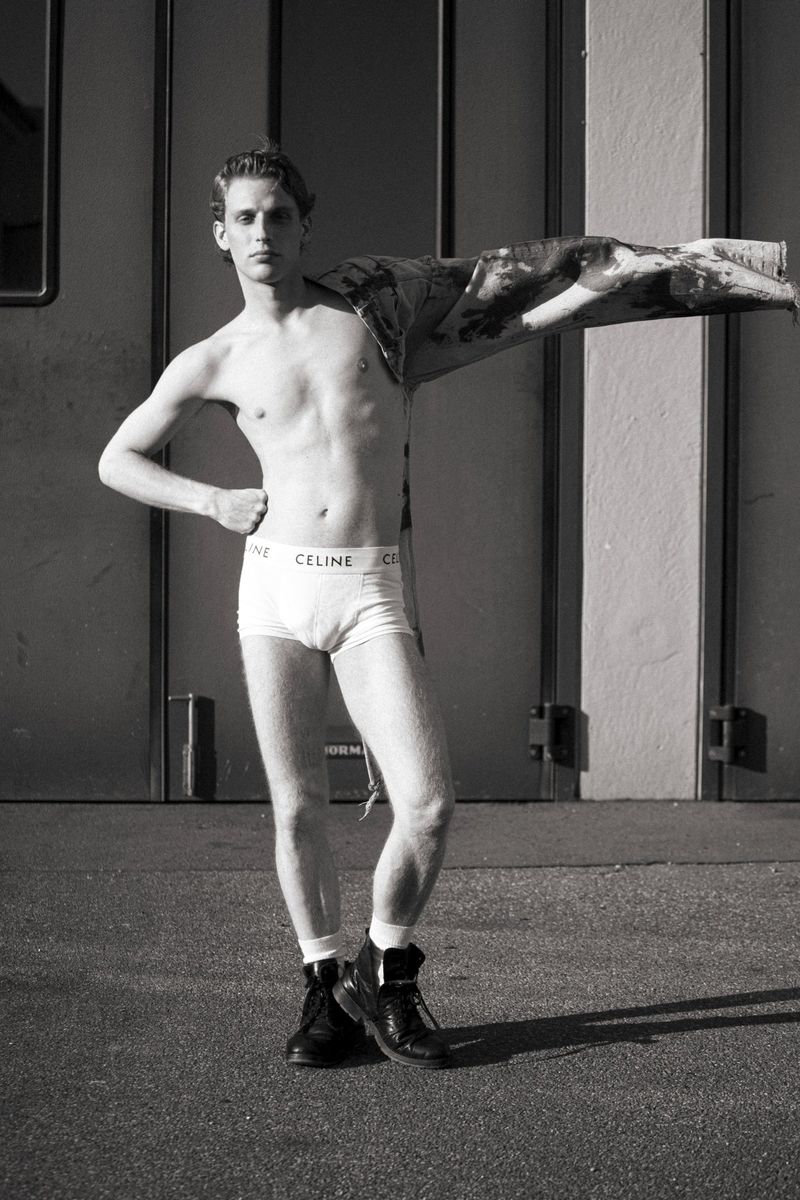 Black-and-white shot of a shirtless adult male posing in briefs and boots outdoors against an industrial wall, with a jacket draped over his shoulder.