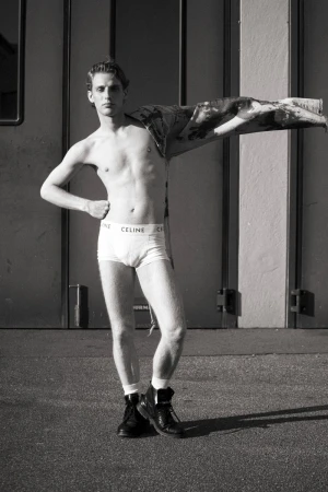 Black-and-white shot of a shirtless adult male posing in briefs and boots outdoors against an industrial wall, with a jacket draped over his shoulder.