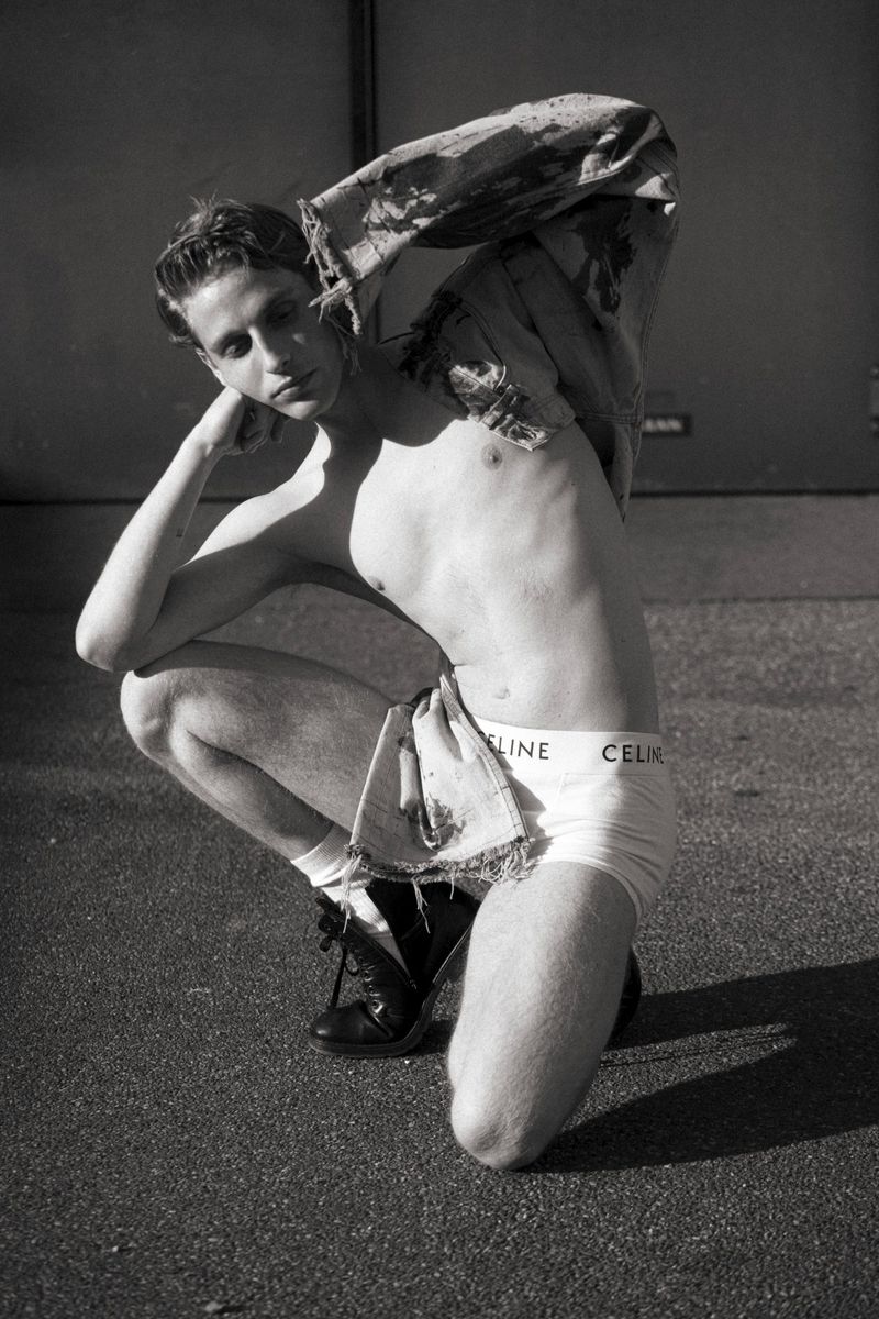 Black and white portrait of a shirtless male model in underwear, squatting outside and posing with his hand near his head.