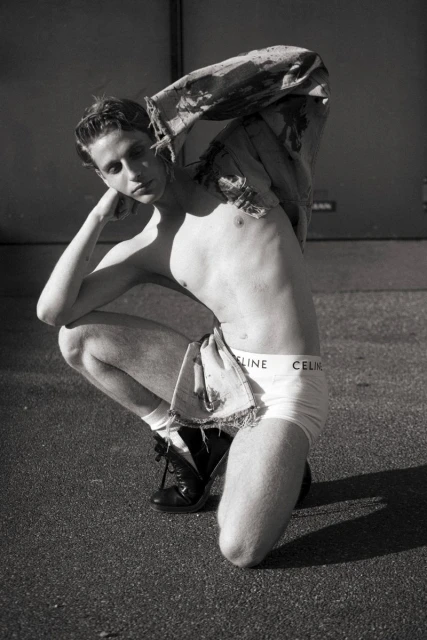 Black and white portrait of a shirtless male model in underwear, squatting outside and posing with his hand near his head.