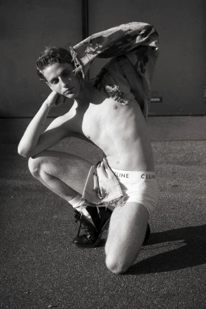 Black and white portrait of a shirtless male model in underwear, squatting outside and posing with his hand near his head.