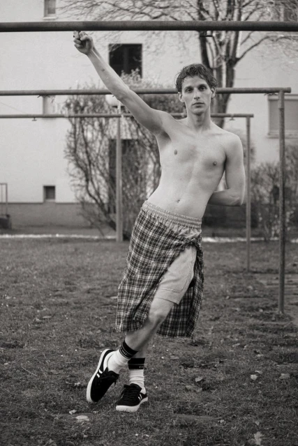 Monochrome photo of a shirtless adult male on playground equipment, wearing a kilt, socks, and sneakers, with an urban backdrop.