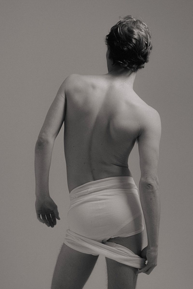 Monochrome portrait from behind of a nude male holding a cloth, with short brown hair, facing away from the viewer in a neutral setting.