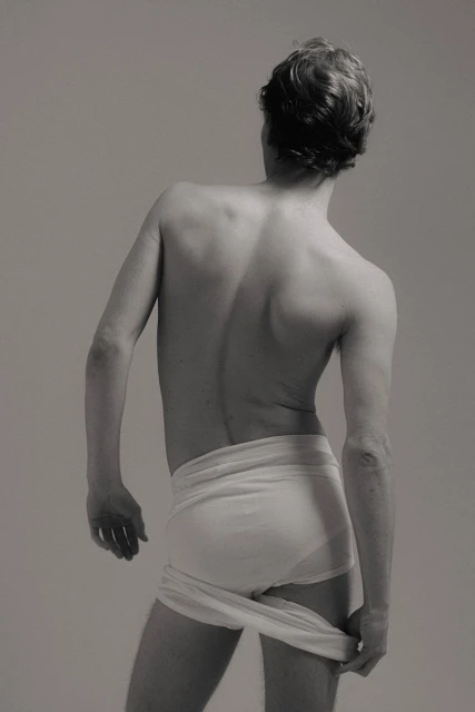 Monochrome portrait from behind of a nude male holding a cloth, with short brown hair, facing away from the viewer in a neutral setting.