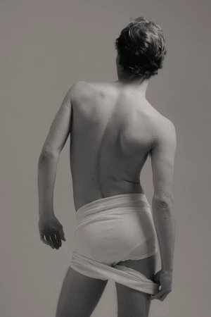 Monochrome portrait from behind of a nude male holding a cloth, with short brown hair, facing away from the viewer in a neutral setting.