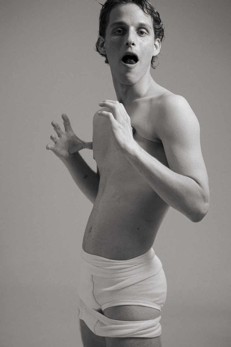 Monochrome studio shot of a fair-skinned, shirtless adult man wearing white underwear, mouth open, and hands raised in a startled expression.