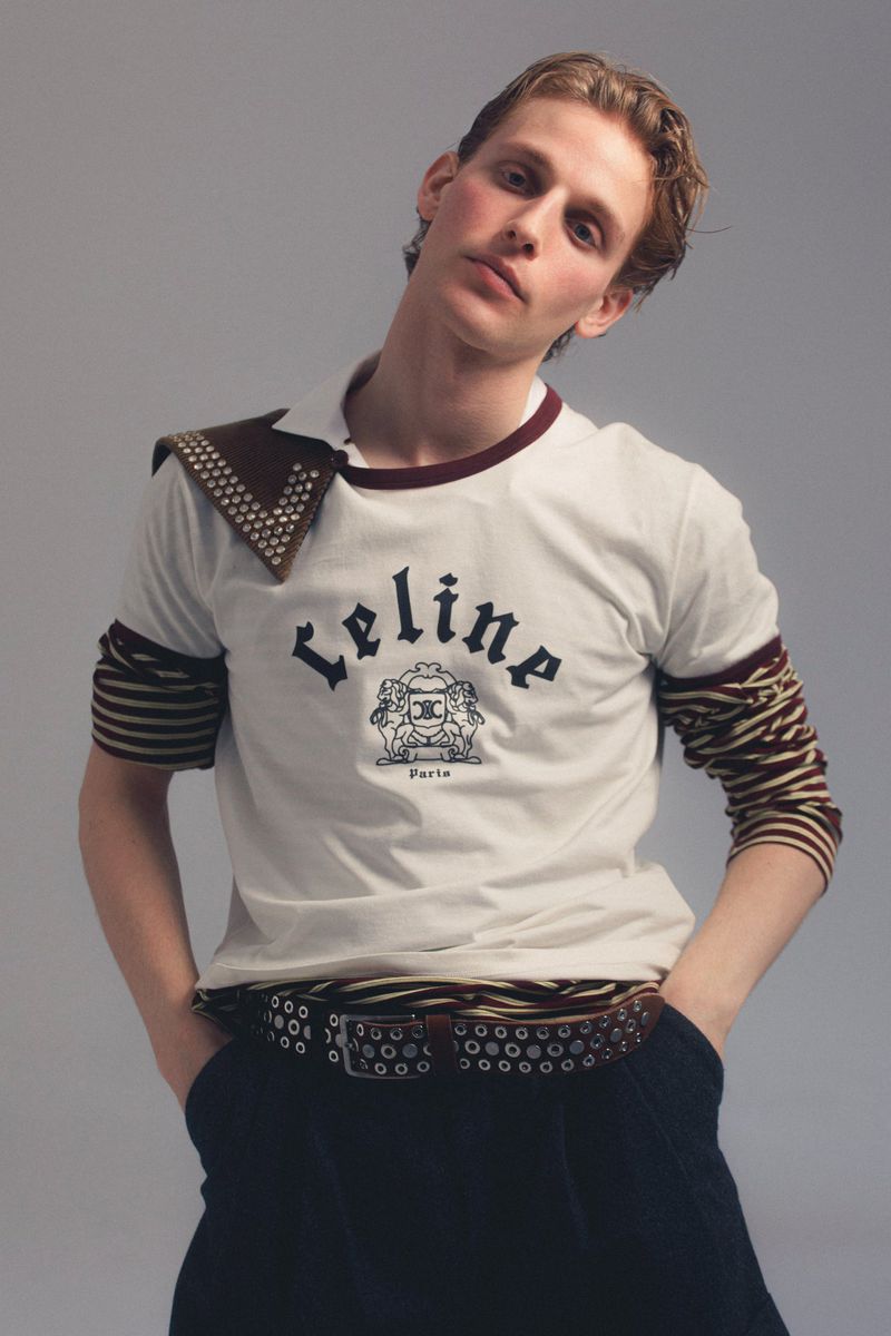Young male model poses in a Celine t-shirt, dark pants, and a studded belt against a neutral backdrop in a studio shot.