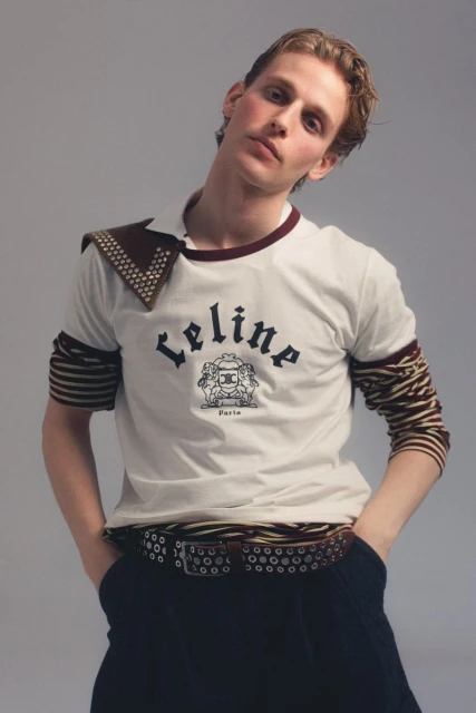 Young male model poses in a Celine t-shirt, dark pants, and a studded belt against a neutral backdrop in a studio shot.