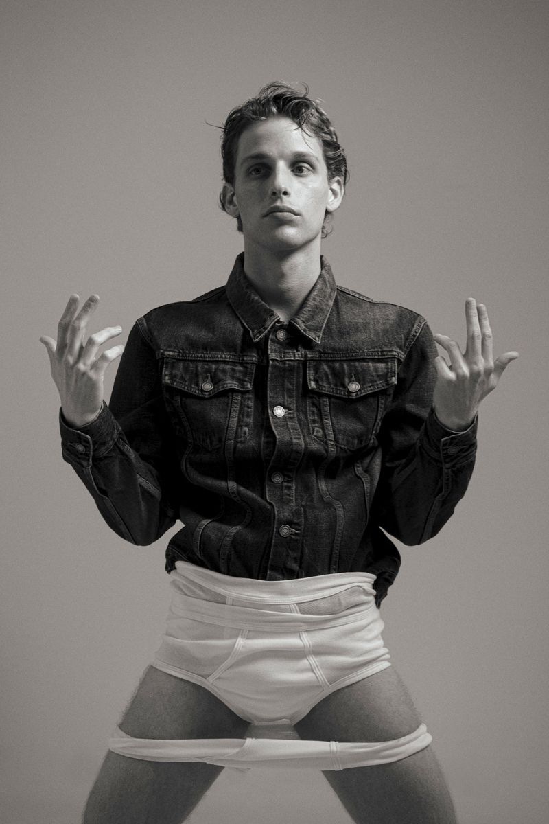 Monochrome portrait of a young adult male in a denim jacket and white underwear, posing in a photo studio against a grey background.