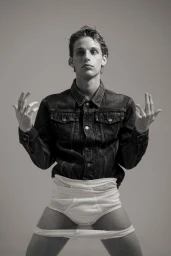 Monochrome portrait of a young man wearing a dark denim jacket and white underwear against a plain grey background