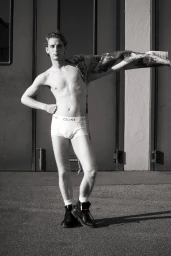 Man wearing white boxer briefs and boots holding up a jacket against an industrial backdrop